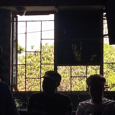 The image shows a dimly lit interior in Kinoo ward, Kenya, with two or three individuals partially visible against a large, multi-paned window. Lush green trees and foliage are visible outside the window, suggesting a verdant environment. The figures appear to be silhouetted, making details about their clothing and features difficult to discern, but they seem to be wearing hats. The overall scene suggests an informal setting, possibly a cafe or gathering space, although specifics are obscured by the low light.