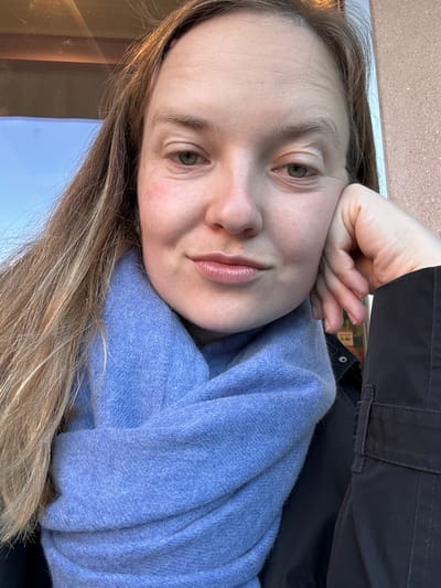A fair-skinned female individual is depicted in a close-up, chest-up shot in Longone al Segrino, Italy. She has long, light brown hair parted on the viewer's left, extending down her shoulders. Her eyes are greenish-hazel, and a slight smile is present on her lips, which have a natural color and sheen. Visible forehead wrinkles and a subtle redness on her left cheek are present. She is wearing a thick, light blue knitted scarf wrapped around her neck, and a dark-colored jacket or coat is visible on her right side, revealing a snap button and part of a utility pocket. Her right hand is raised, with fingers curled, supporting her jawline. The background, out of focus, shows a warm-toned, textured wall or building surface. To the viewer's left, a reflection of a blue sky is visible, likely from a window. Hints of indistinct colorful elements are visible in the background to the right.