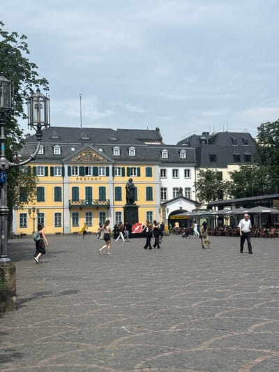 This image captures a view of the market square (Marktplatz) in Bonn, Germany. The central focus is the yellow building labeled "POSTAMT," indicating a post office. In front of the Postamt, a statue of a man stands on a pedestal. The square is paved with cobblestones, and several people are walking around. To the right of the Postamt, there is an outdoor cafe with tables and umbrellas. In the background, other buildings and trees are visible under a cloudy sky. The scene appears to be a public space with commercial activity and pedestrian traffic.