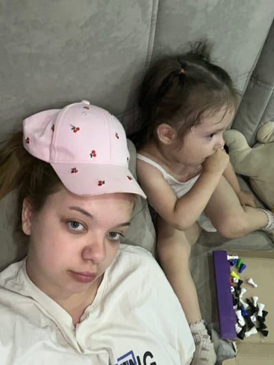 The image is a selfie taken indoors, likely on a couch, with two subjects. The foreground features a young woman wearing a pink baseball cap with a cherry pattern and a white t-shirt. Her gaze is directed towards the camera. To her right and slightly behind her, a young child is sitting and looking away from the camera, with their hand near their mouth. The child is wearing a white tank top and patterned socks. Scattered on the surface in front of the child are pieces from a board game, appearing to be chess pieces, along with a purple game component. The background is a neutral-colored, textured surface, possibly upholstery. There is no discernible information in the image that suggests a location in Gorodets, Russia.