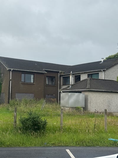 The image shows an overcast day in Bleary, United Kingdom, with a focus on two adjacent houses that appear to be abandoned or under renovation. The house on the left has a dark brown brick facade and boarded-up lower windows. The house on the right is constructed of a lighter-colored, textured material and features several windows, some of which are dark or appear to be damaged. Both houses have dark tiled roofs and white guttering.

The foreground is dominated by overgrown grass and weeds, with a wooden fence and wire running across it. A small, bushy green plant is situated near the fence. In the background, beyond the houses, some trees are visible, suggesting a semi-rural or suburban setting. The sky is a uniform grey, indicating cloudy weather. There are no people or animals visible in the image. The overall impression is one of neglect and disuse.