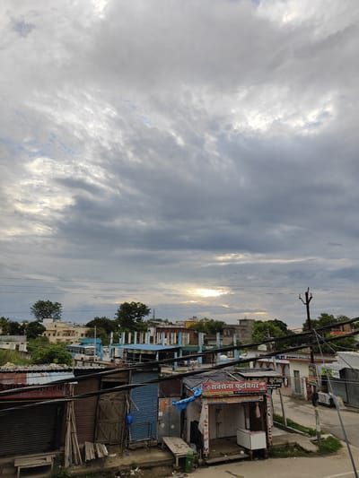 This image captures a view of a street in Sitarganj, India, under a cloudy sky. The foreground is dominated by several small shops with corrugated metal roofs and blue and brown shutters. Some shops have signs in Hindi, one of which reads "SAKSENA FOOTWEAR." Wooden planks and a bench are visible outside one of the shops. Power lines crisscross the scene, supported by wooden and metal poles. In the background, there are buildings, including what appears to be a multi-story structure with blue trim and possibly under construction. Trees and a bus are also visible among the buildings. The sky is filled with dramatic, textured clouds, with a patch of sunlight breaking through in the upper center of the image, casting a glow. Several small birds are visible in flight against the sky.