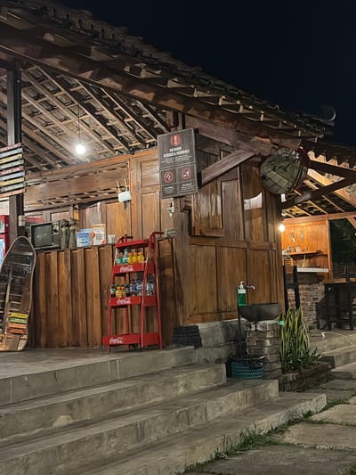 The image shows a wooden structure with a thatched roof, possibly a restaurant or stall, at night in Campurejo, Indonesia. The structure has multiple levels and is made of natural wood. A red Coca-Cola display rack with several bottles of beverages is prominently placed in front of the wooden wall. Several signs are visible, including a warning sign with text in Indonesian and a QR code labeled "KRITIK & SARAN". A mirror reflecting the interior of the establishment is mounted on the wall. Steps leading up to the structure are made of concrete. Greenery is visible in the foreground and to the right of the structure. The lighting suggests it is evening or nighttime. No people are clearly visible in the image.