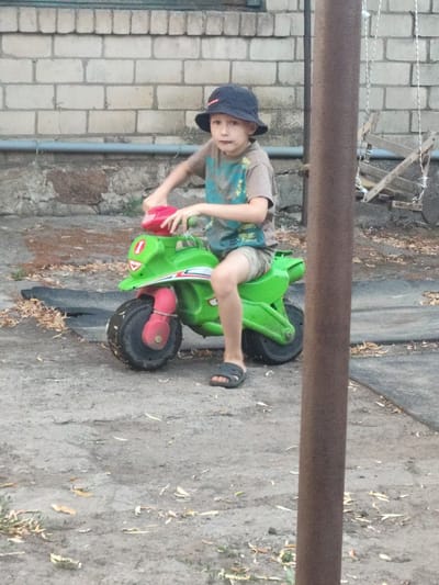 In Saksahan, Ukraine, a young boy wearing a dark bucket hat and a grey t-shirt with a colorful graphic rides a bright green toy motorcycle. He is positioned on a paved surface with scattered leaves. The background features a brick wall, a metal pole, and a detached swing set. The boy's feet are clad in black sandals. The toy motorcycle has red accents on the handlebars and front wheel.
