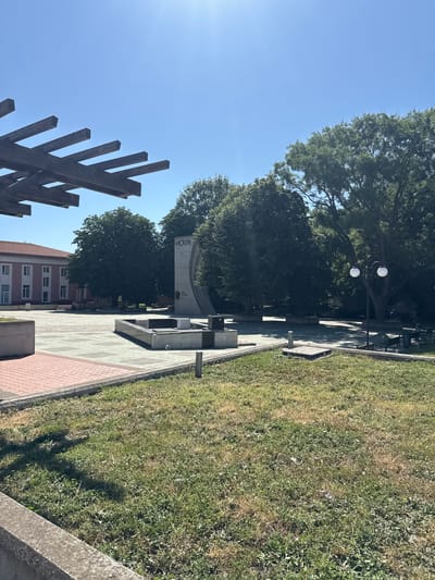 The image shows a public square in Shabla, Bulgaria, on a sunny day. In the foreground, there is a patch of dry grass and a concrete barrier. A wooden pergola extends from the left side of the frame. The middle ground features a paved square with a monument in the center. The monument is a tall, grey structure with curved elements and text inscribed on it, appearing to be Cyrillic. Beside the monument is a low, water feature or fountain. Beyond the square, there are mature trees and a building with a pink facade and white windows. A street lamp with two globes stands near the trees. The sky is clear and blue. The scene depicts a quiet, outdoor public space.