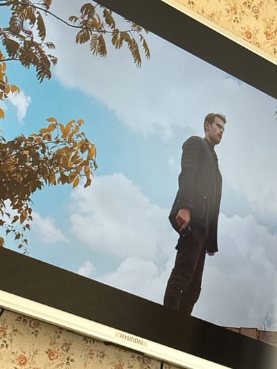 The image depicts a man wearing a dark jacket and trousers, seen from a low angle, appearing to float against a blue sky with scattered clouds. Some trees with autumnal-colored leaves are visible in the lower part of the image, suggesting an outdoor setting. The picture appears to be displayed on a screen or monitor, possibly a Hyundai model as a logo is visible, with a floral-patterned surface in the background. The location is not discernible from the image.