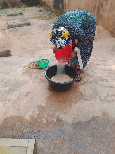 The scene is set outdoors in Ufuma, Nigeria. A person, wearing a multi-colored patterned head covering (blue, yellow, white, black), a dark blue floral patterned dress with white side trim, and dark sandals, is bent forward. The individual is pouring a stream of whitish liquid and yellowish grains from a red plastic basin into a larger black plastic basin positioned on the ground. The black basin contains yellowish grains partially submerged in the liquid. To the left, a small woven tray holds a green plastic colander and a quantity of dry, brownish granular material. The ground consists of uneven concrete or compacted earth, displaying numerous wet patches and water stains, particularly around the black basin. In the background, a light brown wall is visible, with sparse green vegetation behind it. Two square, raised concrete structures are present in the left background.