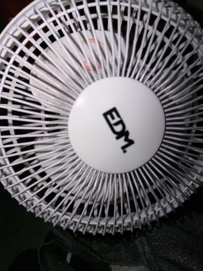 An extreme close-up, slightly tilted, overhead shot features a white electric fan. The fan's grille is a dense network of white, thin, vertical bars interspersed with a few horizontal bars, creating a cage-like effect. In the center of the fan, a circular, white hub is visible with the black logo "EDM." embossed on it. The fan appears to be dusty. The background is dark, suggesting the fan is not in direct light. There are no people or animals present in the frame. The image does not provide any information about its location in Torremolinos, Spain.