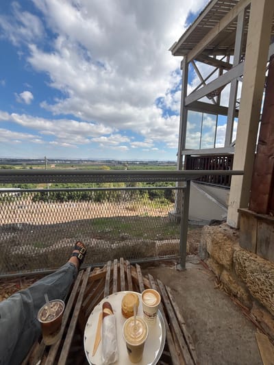 The image presents an outdoor patio scene in Kfar Vitkin, Israel, offering a wide view of an agricultural landscape under a blue sky with scattered white clouds. In the foreground, a person's left leg, clad in blue jeans and a black two-buckle sandal, is partially visible, resting on a wooden slatted surface. On this surface, a white round tray holds a white paper cup with a latte displaying heart-shaped latte art, a clear plastic cup containing a blended light-colored beverage, a round light-brown pastry, and a long item wrapped in white paper with a small wooden knife and spoon. A separate clear plastic cup with an iced coffee rests near the person's leg on the same wooden surface. A dark grey metal balcony railing with two horizontal bars and diamond mesh infill runs horizontally across the midground. Beyond the railing, the ground slopes downward with sparse vegetation, leading to vast green agricultural fields that extend to the distant horizon, where faint hills or mountains are discernible. On the right side, a modern building structure with prominent wooden beams and a large glass panel reflecting the sky is visible. Large, irregular tan stones form a part of the building's foundation next to the concrete patio floor.