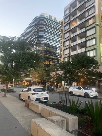 A street scene in Brisbane, Australia, captures modern architecture and urban life at dusk. The foreground features a paved sidewalk with concrete blocks and planters containing green foliage. A white Toyota Corolla hatchback is parked on the street, facing away from the camera, with its license plate clearly visible. Next to it, a white Tesla Model Y is parked, also facing away. A black bicycle rider is visible in the background on the street.
Dominating the background are two prominent buildings. To the left, a contemporary office building with a curved facade and numerous horizontal windows reflects the soft evening light. To the right, a multi-story apartment building displays a grid of balconies with glass railings and a mix of lit and unlit windows. The street is lined with trees, some adorned with decorative string lights, hinting at an evening ambiance. The sky above is a clear, pale blue, indicative of twilight. The overall impression is one of an urban environment transitioning from day to night.