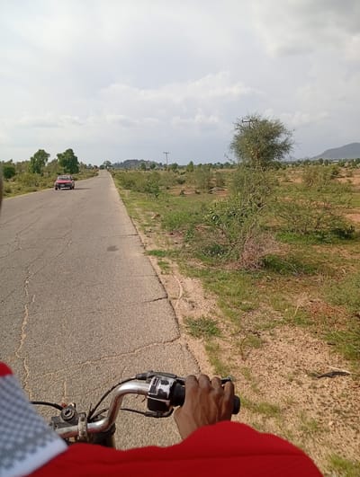 The image shows a view from the perspective of someone riding a motorcycle on a road in Shuwa, Nigeria. The road is asphalt and appears to be in moderate condition, with visible cracks. To the left of the road, a red car is visible in the distance, driving in the same direction. The landscape on either side of the road is rural, characterized by sparse vegetation, scattered trees, and dry, sandy soil. In the background, hills are visible, and the sky is overcast with clouds. A power pole with wires is also present on the right side of the road. The handlebars and a portion of the rider's arm and red clothing are in the foreground.