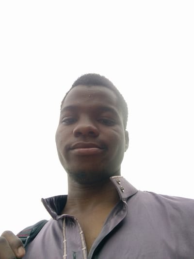A low-angle close-up selfie of a dark-skinned man with short, dark hair and a slight beard. He is wearing a lavender shirt with a high collar and two visible silver buttons. Part of a backpack strap is visible over his left shoulder. The background is a bright, overcast sky. No specific details indicate the location is Eleweran, Nigeria; this is not confirmed.