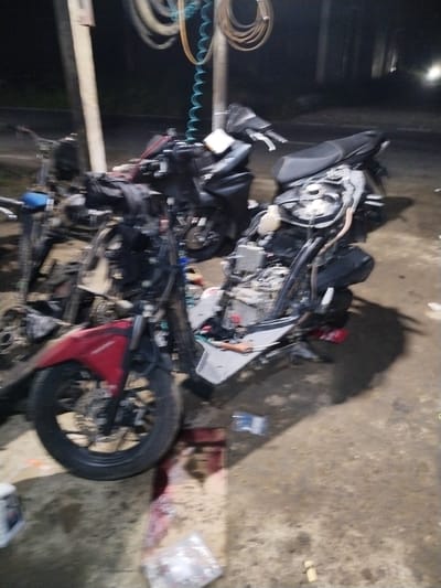 The image depicts an outdoor scene at night in Meunasahpantonlabu, Indonesia, featuring at least two damaged or disassembled scooters. One scooter, primarily red and black, is in the foreground, its front wheel and part of the frame visible. The second scooter, mostly black, is positioned behind and to the left of the first, also appearing to be dismantled or in disrepair. Both scooters show exposed mechanical components. The ground is a dirt or gravel surface, characteristic of an unpaved area. There are no people visible in the image. The lighting suggests artificial light sources, creating shadows and illuminating parts of the scene. Some hoses or cables are visible hanging from a structure on the left, and various debris or parts are scattered around the scooters.