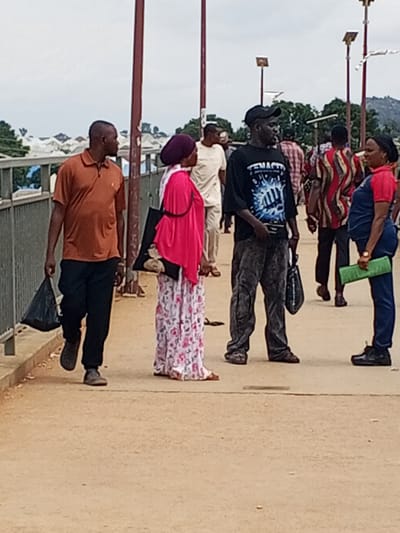 The image shows a group of people on a bridge or walkway. In the foreground, a man in a rust-colored polo shirt and black pants carries a black plastic bag in his left hand. To his right, a woman wearing a pink headscarf and a long, floral-patterned dress holds a black bag. Next to her, a man in a black t-shirt with white text and dark, baggy pants holds a black bag. To the right of this man, a woman in a blue overall and a red t-shirt stands with a green object, possibly a mat or a book, under her arm. Other people are visible in the background, some with colorful clothing. The setting appears to be outdoors, with trees and buildings in the distance, suggesting an urban or semi-urban environment. The sky is overcast, indicating a cloudy day. The overall atmosphere seems to be one of everyday activity, with people moving about. The visible text on the black t-shirt reads "TENACIT".
