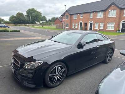 A black Mercedes-Benz E-Class coupe is parked on an asphalt driveway in front of a row of modern brick townhouses in Craigavon, United Kingdom. The car's front passenger side is prominently displayed, showcasing its alloy wheels and headlights. The townhouses feature dark gray tiled roofs and white-framed windows. A Union Jack flag is attached to a lamppost in front of one of the houses. In the background, there are trees and a cloudy sky. A blue car is visible further down the street, and a portion of a silver car is in the foreground, partially obscuring the view of the black Mercedes. The environment appears to be a residential area.