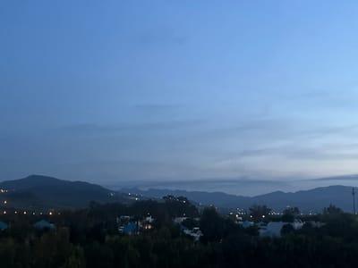 This wide-angle landscape photograph captures a tranquil scene at twilight or night, looking over a settlement nestled within a mountainous region. The sky, dominating the upper half of the image, presents a deep, gradient blue, transitioning to a slightly lighter, hazier blue towards the horizon, with faint, wispy cloud formations.
In the mid-ground and background, multiple layers of dark mountains and hills are silhouetted against the sky, their rugged outlines forming a serene backdrop. Below these mountains, a sprawling settlement is visible, characterized by numerous houses and buildings. Many of these structures feature distinct light blue roofs and white walls, interspersed with dense, dark green trees and vegetation.
A prominent feature of the scene is the scattering of small, bright, yellow-orange lights, indicating human habitation. These lights are distributed throughout the settlement, some forming a clear horizontal line in the mid-left, possibly delineating a road or a residential area. Other lights twinkle sporadically across the hillsides, suggesting homes or infrastructure extending into the higher elevations. On the far right, a slender, dark tower-like structure stands against the twilight sky.
The overall impression is one of peacefulness and quiet, with no discernible human activity. The setting, with its distinct architecture and mountainous terrain, aligns with the provided location context of Fnideq, Morocco.
