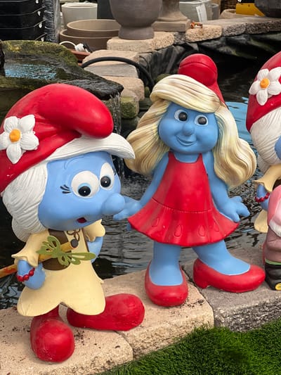 This image shows a collection of Smurf figurines, likely garden ornaments, displayed near a water feature. One figurine is Smurfette in a red dress, while another is a male Smurf with a white beard, red hat with a white flower, and a musical instrument. The figurines are positioned near a stone-like structure, with a glimpse of green grass visible in the background. The scene appears to be outdoors, possibly in a garden or a retail setting selling garden decorations. There is no information present to determine the location as Zvulun Regional Council, Israel.