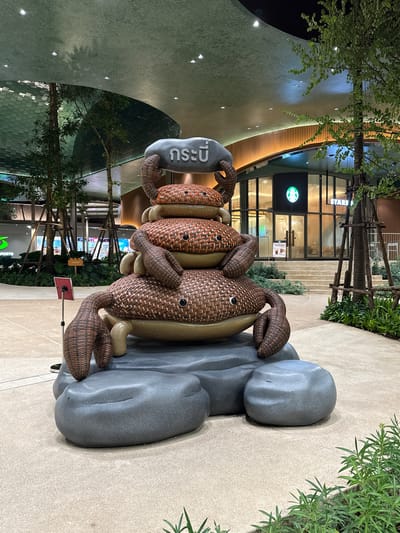 The image shows an outdoor public space at night, featuring a large, stacked crab sculpture as the central focus. The sculpture is made of woven material in shades of brown and has a stone-like base. Above the sculpture, a grey stone with Thai lettering is visible. In the background, a Starbucks store with its iconic green logo and illuminated interior is seen, with stairs leading up to its entrance. Several trees with visible branch structures are present, along with modern architecture featuring curved, green ceilings and wooden paneling. The ground is paved with light-colored tiles. There are no people visible in the image. The setting appears to be a modern commercial or recreational area.