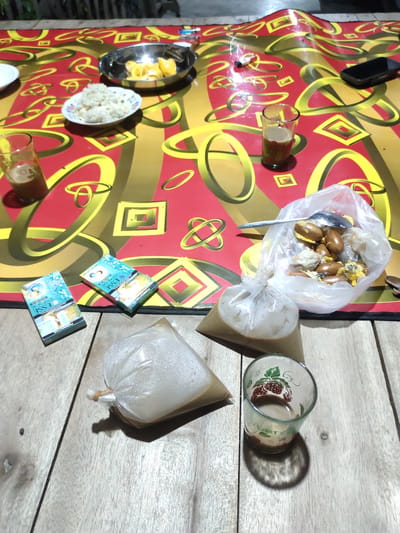 An overhead, dimly lit, eye-level shot captures a casual gathering outdoors on a wooden table covered with a red and yellow patterned tablecloth. In the background, the dark silhouettes of people and possibly structures are visible. The foreground is dominated by food and drinks. A small silver bowl holds pieces of yellow fruit, possibly mango or jackfruit, next to a small white dish filled with white rice. Three glasses are present: one partially filled with a brown liquid, another empty but decorated with a strawberry pattern, and a third, taller glass containing a brown beverage. Two packs of "Esse" brand cigarettes, blue and white, are placed near the glasses. A clear plastic bag holds more of the yellow fruit, with a spoon resting on top. Another clear plastic bag, tied with a rubber band, contains a pale beige liquid, as does a conical plastic bag filled with a darker beige liquid. A black smartphone lies on the tablecloth in the upper right corner. The scene suggests a relaxed meal or break.