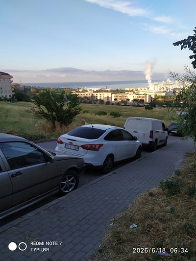 The image captures a view of a street in Avcılar, Turkey, during daylight hours. In the foreground, two cars are parked on a brick-paved sidewalk. The car on the left is a silver sedan, and the car on the right is a white Ford Focus. Further down the street, a white van is parked. Beyond the parked vehicles, a grassy slope rises, dotted with dry vegetation and a few trees. A person in a light-colored shirt is partially visible standing near the white car on the slope. In the background, a cityscape unfolds with buildings, including a multi-story apartment building on the left. Across a body of water, industrial buildings with smoke plumes rising from stacks are visible, suggesting a port or industrial area. The sky is clear with some wispy clouds. The image also contains timestamps and text overlays: "REDMI NOTE 7", "ТУРЦИЯ", and "2026/6/18 06:34".