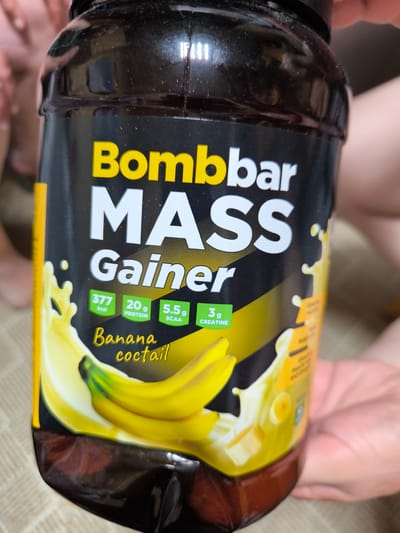 The image shows a close-up of a black plastic tub of "Bombbar MASS Gainer" protein supplement. The tub has a black label with yellow and white text and graphics. The prominent text on the label includes "Bombbar MASS Gainer" in large white letters, with "Bombbar" in yellow. Below "MASS Gainer" are smaller green boxes with white text indicating nutritional information: "377 kcal", "20g PROTEIN", "5.5g BCAA", and "3g CREATINE". The flavor is identified as "Banana cocktail" in yellow cursive writing. A graphic of bananas and splashing liquid, depicting a banana shake, is visible on the right side of the tub. The background is blurred, showing what appears to be people's hands and possibly clothing, suggesting the photo was taken indoors. The lighting appears to be artificial, and the overall mood is neutral. There are no specific indicators of time of day, weather, or a distinct location within the image itself, beyond the label text. The context of "Gal’chino, Russia" is external information.
