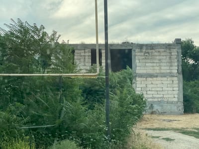 The media file depicts an unfinished or abandoned building constructed from white cinder blocks. The building is rectangular with open doorways and appears to be a single-story structure. It is partially obscured by lush green foliage, including dense bushes and trees, suggesting an overgrown or natural setting. Two vertical poles, one light-colored and one dark, are positioned in front of the building, one closer to the viewer than the other. A horizontal pipe runs across the foreground, further separating the viewer from the building.
The sky is overcast with gray clouds, indicating a potentially cloudy or dull day. The ground around the building is covered in dry grass and some scattered gravel or dirt. There is graffiti visible on the lower portion of the building's walls, with some letters and symbols. The overall impression is one of disuse and decay, with nature encroaching on the man-made structure. The time of day is unclear due to the overcast weather, but it appears to be daytime. The location is identified as Congaz, Moldova. No people or specific activities are depicted in the image.