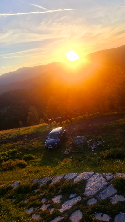 The image depicts a scenic sunset over a mountainous landscape, with a car parked on a grassy slope and several horses grazing nearby. The sun is low on the horizon, casting a warm, golden glow across the scene. The sky is a vibrant mix of orange, yellow, and blue, with wispy clouds streaking across it. In the foreground, a dark-colored car is parked on the grass. Several horses are grazing on a dirt path behind the car, their silhouettes dark against the bright light of the sunset. The surrounding landscape is hilly and covered in trees, with a prominent mountain range visible in the distance. The overall impression is one of natural beauty and tranquility. The location of Simeonovets, Bulgaria, is not explicitly visible in the image, but the mountainous terrain suggests a rural or natural setting.