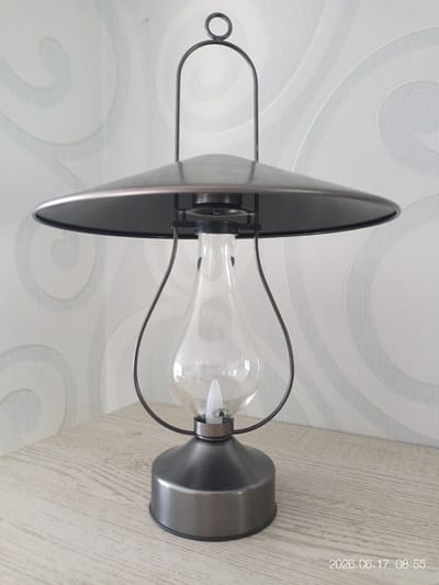 The image shows a decorative lantern with a dark metal finish and a clear glass globe, resting on a light-colored wooden surface. A thin, dark metal handle arches over the top of the lantern, with a small loop at its apex. The lantern is positioned in front of a wall with a subtle, light-colored, swirled wallpaper pattern. The overall impression is of an indoor setting, possibly a shelf or table. The date and time stamp "2026.06.17 08:55" is visible in the lower right corner. There are no people or discernible actions occurring in the scene. The location is not identifiable from the visual information provided; therefore, it cannot be confirmed as Beringen, Belgium.