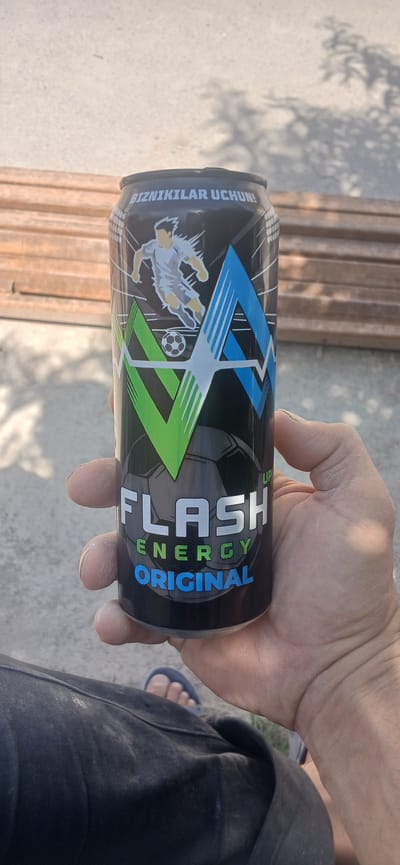 In the city of Daliguzar, Uzbekistan, a person's hand is holding a black aluminum can of "FLASH ENERGY ORIGINAL" energy drink. The can features a graphic of a soccer player in action, a soccer ball, and abstract shapes in green and blue. The background consists of a concrete surface and wooden planks. The person is wearing dark pants and a sandal on their visible foot. The scene is illuminated by natural light, casting shadows from surrounding foliage onto the ground.