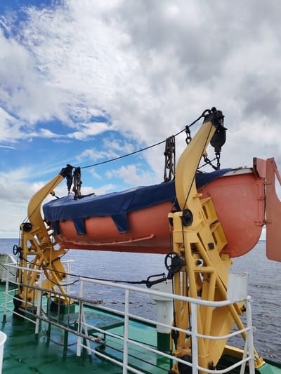 The image shows a lifeboat suspended on a davit system aboard a ship. The lifeboat, orange in color, is covered with a blue tarp. The davit system is painted yellow and is a complex arrangement of beams, pulleys, and ropes. The ship's deck, visible in the foreground, is painted green and has white railings. The sea is visible in the background, with a cloudy sky above. The image does not contain any information that allows for its localization in Kostyaevka, Russia. There are no people visible in the image.