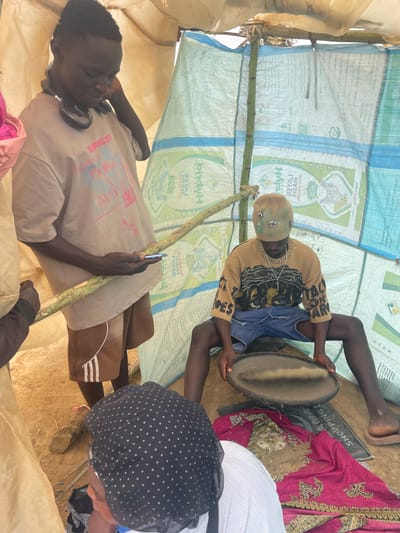 Captured indoors, the image presents a group of individuals within a makeshift shelter constructed from repurposed fertilizer bags, bearing the brand "Hybrid."
Centered, a seated person, adorned with a decorated beanie and a patterned top, holds a circular object, possibly a drum or tray. To the right, partial views of another person with a polka-dotted head covering and a piece of pink fabric are seen.
On the left, a man in a graphic t-shirt and headphones holds a slender, green cane or stick, also interacting with a phone.
The shelter's interior reveals a dirt ground and minimal furnishings, adding to the sense of poverty. A "SHOPPING AMA" sign lies near the seated person. The image appears to be a candid snapshot of everyday life in Maiyanga, Nigeria.