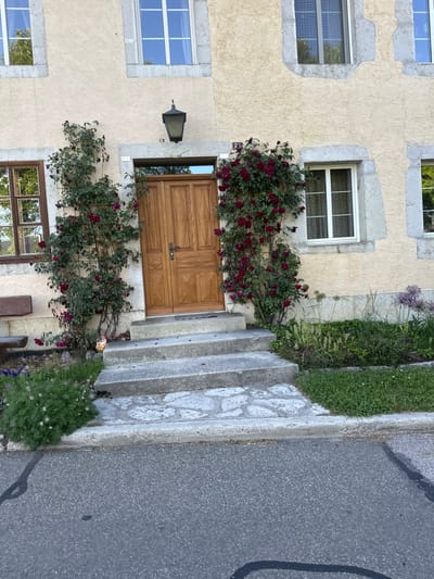 The image shows the exterior of a house in Le Sentier, Switzerland. The house has a light yellow stucco facade with grey stone trim around the windows and doors. A wooden door is centered on the facade, accessed by a set of concrete stairs with irregular stone pavers on the landing. Red roses are climbing the wall on either side of the door, with a green bush and a small garden gnome at the base of the left rose bush. There are several windows visible, some with white frames and others with dark frames. A street runs in front of the house, with a curb and asphalt pavement. The overall scene is one of a residential property with a well-maintained garden.