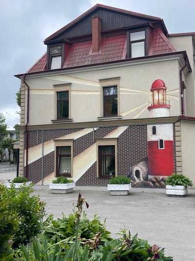 The image shows the side of a building with a mural of a lighthouse painted on it. The building has a red tiled roof and several windows. In front of the building are three white planters filled with green plants. The mural depicts a red and white lighthouse with a beam of light emanating from its top. The lower part of the mural is designed to look like brickwork. The scene appears to be on a cloudy day, and there is a paved area in front of the building. The location is identified as Pionersky, Russia.