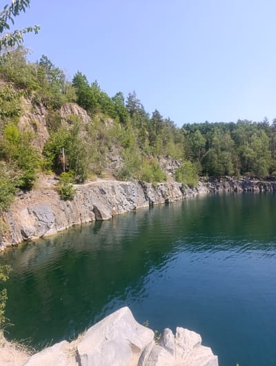 In Výkleky, Czechia, a quarry has been transformed into a scenic lake. Steep, rocky cliffs line the left side of the image, with lush green trees and vegetation growing on their ledges and crest. The water of the quarry is a deep, dark green, reflecting the surrounding foliage and the clear blue sky. A lone individual, wearing orange shorts, stands at the edge of the water near the base of the cliff, seemingly preparing to enter the lake or having just emerged. The foreground shows the rough texture of large, light-colored rocks, suggesting the viewer is standing on another part of the quarry's edge. The overall impression is a tranquil, natural setting ideal for recreation.