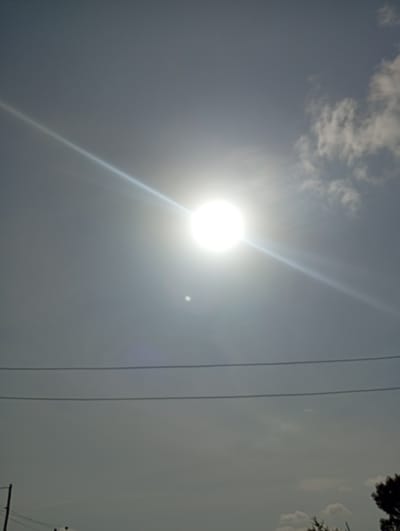The image shows a bright sun in a slightly hazy sky, casting a beam of light downwards. Two power lines are visible horizontally across the frame. In the lower portion of the image, a telephone pole and some trees are partially visible, suggesting an outdoor setting. The overall impression is of a sunny day. There are no people or specific identifiable landmarks to pinpoint the location within Tilde, Nigeria.