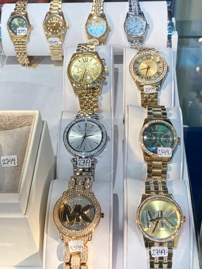 The image shows a display of Michael Kors watches in a store located in Herning, Denmark. The watches are arranged in rows on white stands within a glass display case. The watches are predominantly gold-toned with some silver-toned options. Many of them feature sparkling embellishments such as rhinestones or diamonds around the watch face and on the bands. Price tags are visible on some of the watches. The lighting suggests an indoor retail environment. No people are visible in the image.