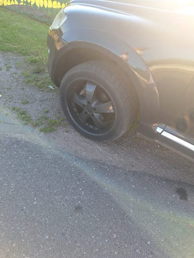 This is a close-up, eye-level, outdoor photograph of the front passenger side wheel and fender of a black SUV. The vehicle is parked on an asphalt surface with a gravel and grass verge to the left. A green metal fence with yellow circular designs is visible in the upper left quadrant, bordering a grassy area. The setting appears to be a residential area in Цімкавіцкі сельскі Савет, Belarus, based on the context. The sun is low and to the right, creating a strong glare and lens flare across the vehicle's body and a faint rainbow effect on the asphalt. The tire is black with a multi-spoke dark grey rim. The fender is black and curves over the wheel well. The vehicle's headlight and part of the hood are visible. There are no people or other distinguishing features in the image.