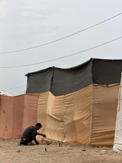 The image, taken in Gaza, shows a man kneeling and working on the ropes securing a makeshift tent or structure. The tent is made of sections of different colored tarpaulins: dark gray, light brown, pale orange, and white. The ground is sandy. Two thin electrical wires run diagonally across the sky. The scene suggests temporary shelter.