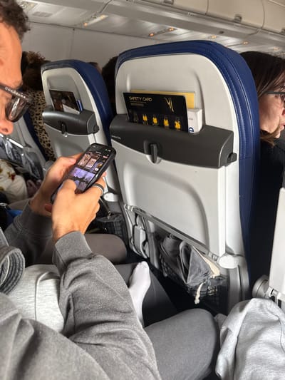 The scene is set inside an aircraft cabin, consistent with either being on the ground at or in flight to/from Lyon-Saint Exupéry Airport (LYS) in Colombier-Saugnieu, France. In the foreground, a person, discernibly male, is seated, wearing a grey hooded sweatshirt, grey sweatpants, and black-framed Gucci glasses. This individual holds a black smartphone horizontally with both hands, actively interacting with the screen, which displays multiple thumbnail images. A black and silver ring is present on the right ring finger, and the right foot, clad in a white sock, is extended. Directly in front of this person is the back of an aircraft economy seat, featuring a white plastic shell with blue trim. Its seat-back pocket contains a black "SAFETY CARD" with gold lettering and graphics of four figures in life vests, alongside a white booklet. Beneath this pocket, a mesh net holds a grey fabric item with a drawstring. To the right, another passenger is partially visible, seen in profile wearing glasses and dark attire. A third passenger, wearing a leopard-print top, is partially visible in the background to the left. The cabin overhead lighting is illuminated.