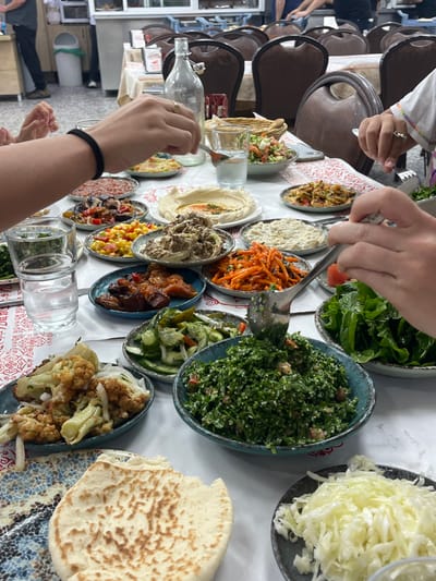 The scene depicts an indoor dining establishment, likely a restaurant, within the Galil Tachton Regional Council, Israel. A large table, covered with a white tablecloth and partially overlaid with a white and red patterned runner, is laden with numerous small, round plates and bowls filled with various mezze-style dishes.
Identifiable dishes include a large bowl of finely chopped tabbouleh being served with a silver spoon by a right hand (adorned with a ring and light pink nail polish), a plate of roasted cauliflower florets, a bowl of shredded white cabbage, a plate of sliced cucumbers and other greens, a bowl of hummus with a central swirl, a plate of corn salad, a bowl of carrot salad, a bowl of a mashed dark dip (possibly eggplant), and several other mixed vegetable and dip preparations. A piece of flatbread rests on a blue and white patterned plate in the foreground.
Multiple clear glass tumblers, some containing water, are distributed across the table, along with a tall, clear glass bottle. At least three individuals are partially visible, represented primarily by their hands and forearms. A left hand, featuring a black elastic wristband and a ring, holds a utensil over a dish.
The background shows more dark brown upholstered dining chairs, suggesting additional tables or seating. Further in the background, a blurred kitchen or service area with light-colored counters, various objects, and indistinct figures of people (possibly staff or other diners) is visible. A white cylindrical trash receptacle is also partially seen. The overall arrangement indicates a communal dining experience.