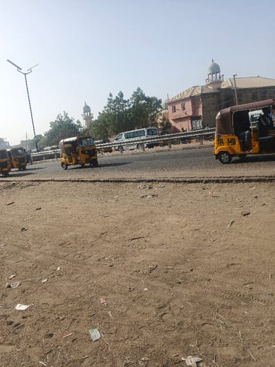 The image portrays an urban street scene in Gobirawa, Nigeria, under a bright, clear sky. The foreground is an unpaved expanse of dry, reddish-brown earth, marked with tire tracks, footprints, and scattered debris including plastic and paper. A paved asphalt road, separated by a concrete curb, traverses the midground. On this road, multiple yellow and brown auto-rickshaws (Keke Napep) are present; one on the right displays "TFA 04B" and contains a driver, while another in the center faces the viewer. To the left, a tall grey street light pole equipped with a dual solar panel array rises. A metal guardrail runs parallel to the road's far side, behind which a white passenger van and several motorcycles are visible. Green trees are interspersed along this section. In the background, a multi-story building with pink and light brown architectural elements is prominent, capped by a large green dome. To its left, a light-colored mosque minaret topped with a smaller dome is discernible.