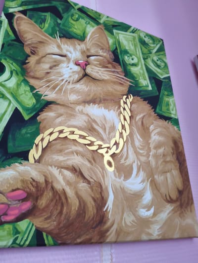 This is a close-up, eye-level, horizontal, medium shot of a painting of a ginger cat wearing a gold chain, lying on a bed of green dollar bills. The cat's eyes are closed, and it appears to be sleeping or resting contentedly. Its nose is pink, and its whiskers are white. The gold chain is thick and has large links. The dollar bills are depicted with the faces of presidents. The background is dark, with the green dollar bills creating a sense of depth. The painting is on a canvas. The lighting is from above, casting soft shadows. The painting is displayed on a light pink wall. The location in the city of Kyiv, Russia is not visible in the image.