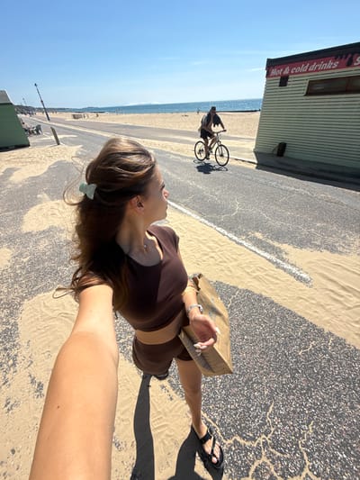 A woman is taking a selfie on a sunny day near the beach in Bournemouth, United Kingdom. She is wearing a brown crop top and shorts, and holding a brown paper bag. She has long brown hair with a light blue hair clip. In the background, a man is riding a bicycle on the road. To the right of the road is a small building with "Hot & cold drinks" written on it. Sand is scattered across the road and pavement. The beach and the sea are visible in the distance under a clear blue sky.
