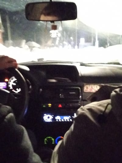 A forensic description of the scene within the city of Урталга, Russia, reveals the interior of a vehicle at night, observed from a rear passenger's perspective. The driver's left hand is visible on the steering wheel, which has an illuminated gauge displaying purple light. To the right, a passenger's torso and arm are partially visible, clad in a light-colored jacket, with the arm resting. The vehicle's dashboard features a central console with an illuminated display showing white circular and humanoid icons, flanked by various indicator lights, including an unlit red hazard triangle and illuminated yellow/orange indicators. Below, two illuminated rotary climate control dials are visible, one green and one blue. A rearview mirror hangs from the ceiling, with a small ornament suspended from it.

Through the windshield, a heavily snow-covered environment is visible, illuminated by bright, diffused light, possibly from vehicle headlights or streetlights. High snowbanks line both sides of the discernible path, suggesting significant snowfall. Distant, blurred figures or objects are barely perceptible beyond the snowbanks. The overall scene depicts a vehicle in operation during winter conditions at night in Урталга.