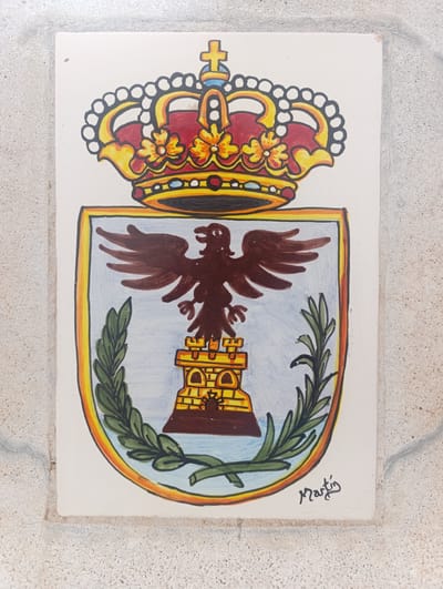The image displays a painted ceramic tile featuring a coat of arms. The coat of arms is topped with a red and yellow crown adorned with white circles and a cross. Below the crown is a shield with a light blue background. Centered on the shield is a brown eagle with outstretched wings. Beneath the eagle is a yellow and brown structure resembling a castle or tower with arches and crenellations. Flanking the castle are green branches with leaves. In the lower right corner of the tile, a signature in dark ink reads "Martín". The tile is set against a textured, light-colored background that appears to be a wall or pavement. This ceramic tile is located on the exterior wall of the "Casa Torre de Cope" in Águilas, Spain.