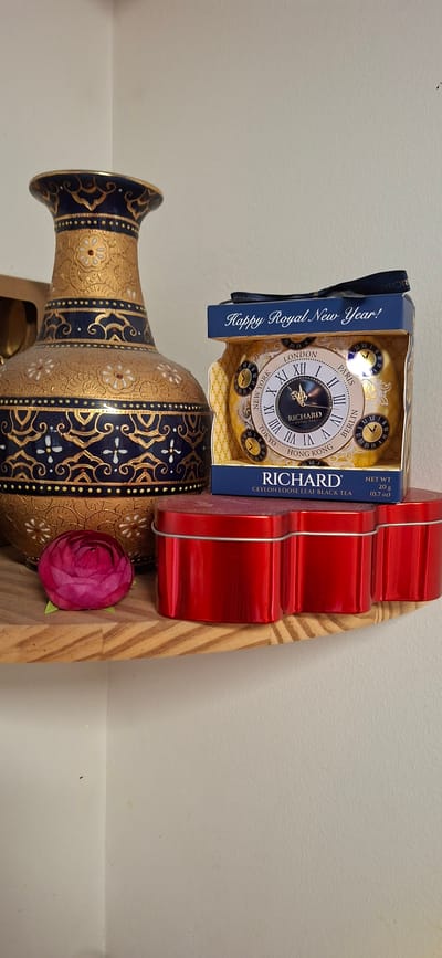 The image shows a collection of decorative items arranged on a shelf. The primary subjects are a tall, ornate vase with intricate gold and navy blue patterns, and a tin of Richard tea. The tea tin is designed to look like a clock face, with Roman numerals and names of cities like London, Paris, and Tokyo around the perimeter. It also features the text "Happy Royal New Year!" and "RICHARD CEYLON LOOSE LEAF BLACK TEA NET WT 20 g (0.7 oz)". Next to the tea tin is a bright red, heart-shaped tin. A single pink artificial rose is placed in front of the vase, adding a touch of color. The shelf appears to be made of light-colored wood with a distinctive striped pattern. The background is a plain, light-colored wall. The lighting suggests an indoor setting, possibly a home or a shop display. There are no people or animals visible in the image. The overall impression is one of curated display, combining ethnic-inspired decor with novelty tea packaging.