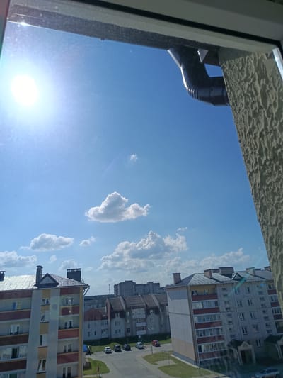 The image shows a view from a window looking out at a bright, sunny day in Smarhon, Belarus. The sky is a vibrant blue with scattered white, fluffy clouds. In the foreground, a dark downspout is visible against a textured, light-colored wall, likely part of the building the photo was taken from.

Below the sky, a collection of apartment buildings fills the middle and background of the scene. The closest building on the left is colorful, with sections of yellow, red, and white. It has many windows, some reflecting the bright sunlight. Further back, more apartment buildings are visible, mostly with gray or beige facades and dark roofs. Several cars are parked on a paved area in front of the buildings, and a single person in red is walking on the sidewalk. The overall setting is a residential urban or suburban area.

The bright sun and clear blue sky suggest it is daytime, likely late morning or early afternoon. The weather appears to be warm and pleasant. The image conveys a sense of calm and ordinary day-to-day life in a European city. There is no visible text in the image.