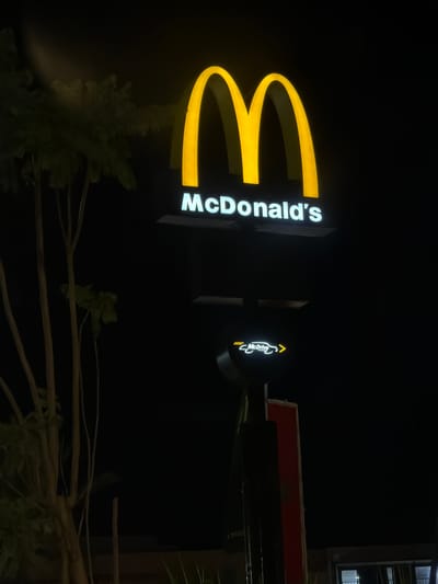A nighttime photograph depicts the illuminated McDonald's sign located in Ouahat Sidi Brahim, Morocco. The sign features the golden arches logo and the name "McDonald's" vertically aligned. A smaller sign indicates "McDrive" with an arrow. Trees and plants are visible in the foreground, partially obscuring the view. The background is dark.