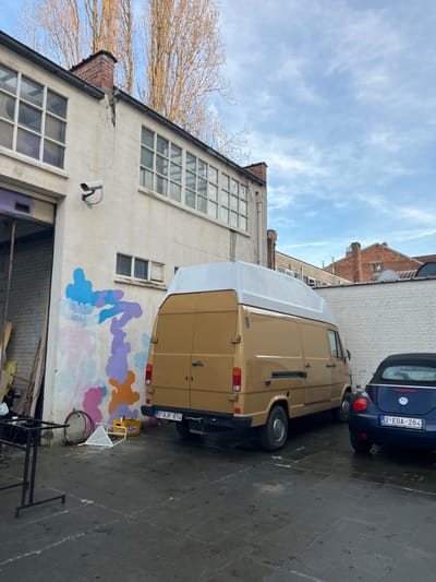 A tan van with a white roof extension is parked on a gray concrete surface next to a blue Volkswagen Beetle. The van's license plate reads "0-AJK-614," and the Beetle's reads "2-EGA-284". Adjacent to the van is a light-colored building with large windows, some broken. A colorful mural, in shades of blue, purple, and orange, adorns the building's wall. A leafless tree is partially visible in the background beneath a cloudy sky.