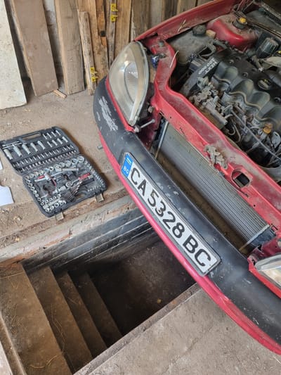 The image depicts the front end of a red car, possibly undergoing repairs, situated near a set of stairs leading to a lower level or basement. The car's license plate reads "CA 5328 BC", indicating it is registered in Bulgaria. A toolbox with various sockets and wrenches lies on the ground next to the car. The location is implied to be Glava, Bulgaria, based on the license plate. The engine bay of the car is exposed, suggesting ongoing maintenance or repair work. The presence of wooden planks near the car may indicate they are being used for support or temporary shoring. The overall environment appears to be a garage or workshop setting.